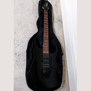 Černá elektrická kytara s tmavým hmatníkem, umístěná v černém poloměkém pouzdře. Na hlavě kytary je bílý nápis Ibanez. Nástroj má dva humbuckery, ovl…