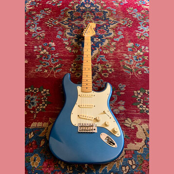 Modrá elektrická kytara typu Stratocaster se světlým javorovým hmatníkem a bílým pickguardem. Na hlavě je vidět nápis Fender Stratocaster. Kytara má…