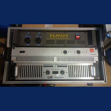 Fotografie zachycuje dva rackové výkonové zesilovače umístěné v transportním kufru. Horní zařízení je černé s nápisem ELMUZ STEREO POWER AMPLIFIER, m…