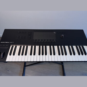 Černý MIDI kontrolér s klávesnicí umístěný na černém stojanu. Na horním panelu je nápis KONTROL S49 MK3, displej a řada ovládacích tlačítek a otočnýc…