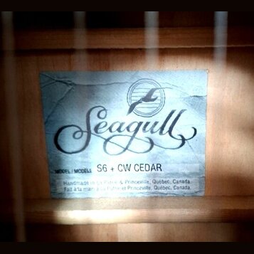 Fotografie znázorňuje štítek uvnitř akustické kytary. Na modrobílém pozadí je černé logo Seagull s vyobrazením ptáka v kruhu. Pod ním je uveden text…