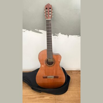 Akustická kytara s vyřezem v korpusu, provedená v přirozené dřevěné barvě. Na hmatníku je vidět štítek s nápisem "Classical Guitar". Nástroj stojí op…