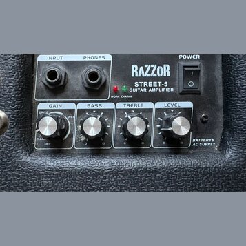 Detailní pohled na ovládací panel kytarového komba s černým texturovaným povrchem. Na panelu je nápis RAZZOR STREET-5 GUITAR AMPLIFIER, vypínač POWER…