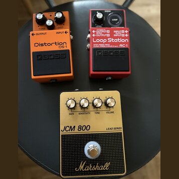 Tři kytarové efekty umístěné na černém podkladu. Vlevo je oranžový pedál s nápisy Distortion DS-1 a BOSS, vpravo červený pedál s nápisy Loop Station…