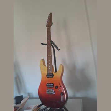 Elektrická kytara s tvarem typu superstrat v přechodovém žluto-červeném odstínu (sunburst). Nástroj má hnědý hmatník, tři černé humbuckery a černý pl…