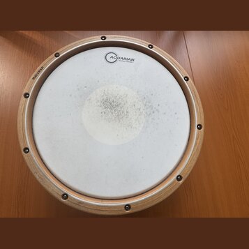 Kruhové bubení s dřevěným rámem a bílou membránou. Na membráně je nápis AQUARIAN TEXTURE COATED a v horní části dřevěného rámu je vyryto HALLVAD. Stř…
