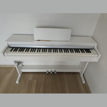 Bílé digitální piano s bílým stojanem a notovým pultem. Na horní liště nad klávesami je umístěno logo YAMAHA. Nástroj má tři st SILVER pedály a někol…