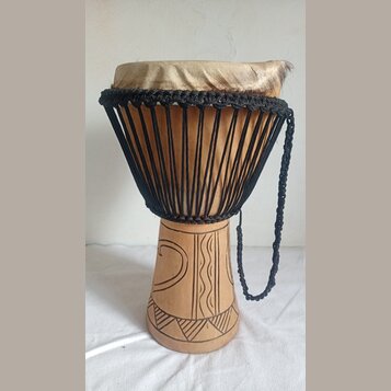 Djembe s tělem z vyřezávaného světlého dřeva a napnutou kůží v horní části. Nástroj je opleten černými provázky, které drží membránu. Spodní část kor…