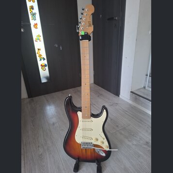 Elektrická kytara typu Stratocaster s javorovým hnedým krkem a tělem v sunburst provedení. Na hlavě krku je svisle napsaný nápis Squier. Kytara má bí…