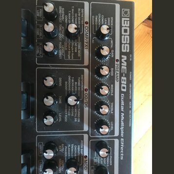 Detailní pohled na ovládací panel efektového procesoru Boss ME-80 Guitar Multiple Effects. Zařízení má černý povrch s bílými nápisy a stříbrnými otoč…