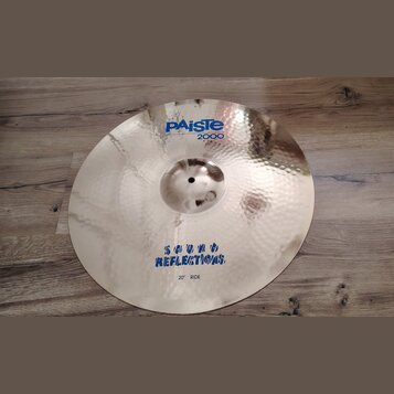 Kovový činel v zlatavé barvě s kovaným, důlkovaným povrchem. Na horní části je modrý nápis PAiSte a pod ním červený nápis 2000. Ve spodní části je mo…