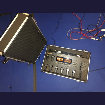 Na fotografii je kytarový procesor Boss GT-100 v bílém hliníkovém kufru s černým polstrováním. Zařízení má digitální displeje, několik ovládacích tla…