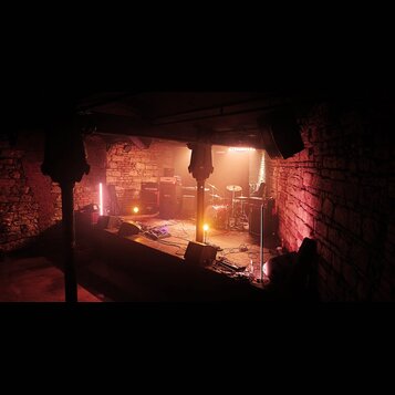 Fotografie zachycuje osvětlené pódium v prostoru s cihlovými stěnami. Na scéně je umístěna bicí souprava, několik kytarových komb a monitorů. V pozad…
