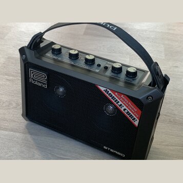 Černý přenosný stereo zesilovač s rukojetí. Na přední mřížce je bílé logo Roland a červenobílý nápis MOBILE CUBE Battery Powered Multi purpose stereo…