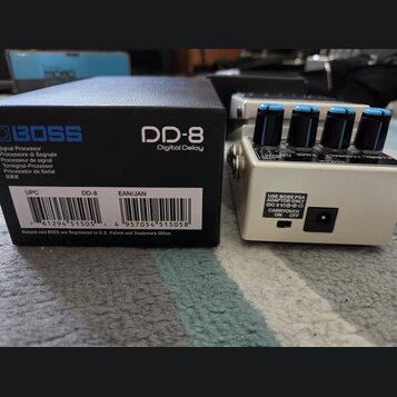 Na fotografii je kytarový efekt Boss DD-8 Digital Delay v bílém provedení s modrými ovládacími knoflíky. Vedle pedálu je umístěna originální černá kr…