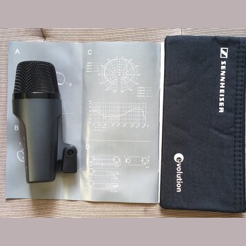 Černý dynamický mikrofon s mřížkou, položený na bílém návodu k použití. Vedle mikrofonu je černý textilní obal s bílým nápisem SENNHEISER a logem zna…