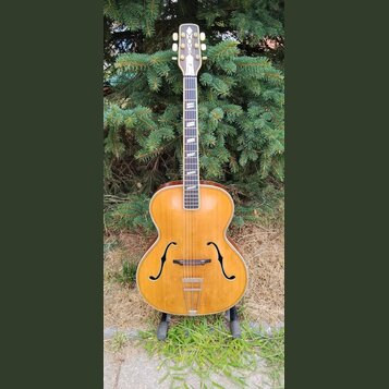 Akustická archtop kytara s přírodním dřevěným povrchem a tmavým hmatníkem. Na hlavě je vidět bílý ornamentální vložkový prvek a šest ladicích kolíčků…