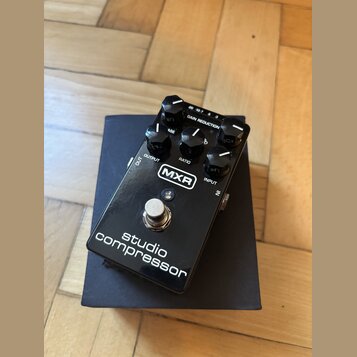 Černý efekt pedál s nápisem MXR a studio compressor. Na horní části jsou čtyři ovládací knoflíky s popisky OUTPUT, RATIO a INPUT, horní regulační prv…