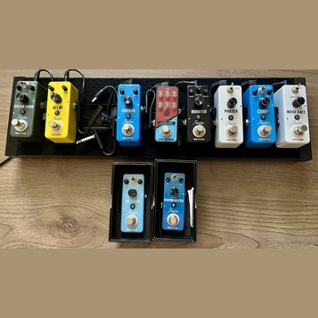 Sada efektů Rewin na černém pedalboardu a dva samostatné pedály v krabičkách. Na boardu jsou zleva doprava pedály v barvách zelené, žluté, modré, čer…