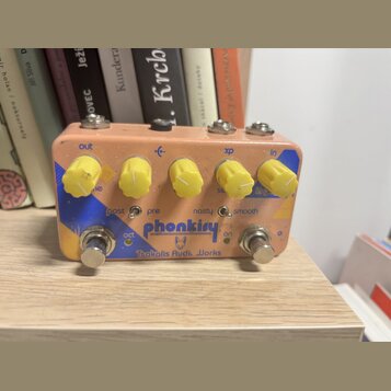 Kytarový efekt pedál v růžovém provedení s žlutými oválnými knoflíky. Na spodní části je modrý nápis phonkiry a pod ním text Tsakalis Audio Works. Na…