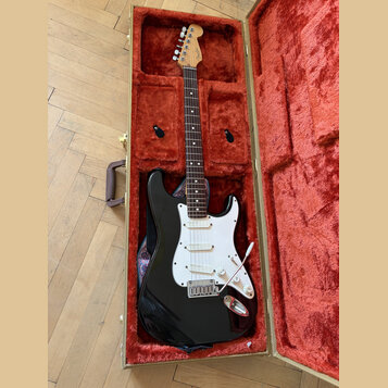 Černá elektrická kytara typu Stratocaster v hnědém tvrdém kufru s červeným plyšovým vnitřkem. Na hlavě hmatníku je vidět nápis Fender. Kytara má bílý…