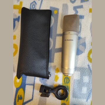 Kondenzátorový mikrofon stříbrné barvy s mřížkou v horní části a nápisem M-AUDIO na těle. Vedle mikrofonu je černý kožený obal se zipem a černý plast…