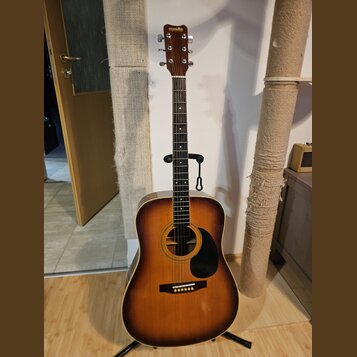 Akustická kytara v barvě sunburst s černým pickguardem a hnědým hmatníkem. Na hlavě je nápis HOHNER. Nástroj stojí na černém stojanu v interiéru. Pov…