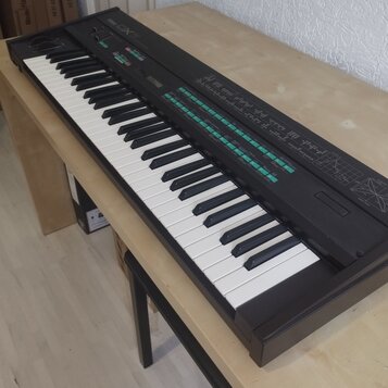 Černý digitální syntezátor s klávesnicí umístěný na světlém dřevěném stole. Na horním panelu je vlevo nápis DX7 a řada barevných tlačítek a ovládacíc…