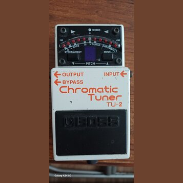 Bílý kytarový ladič s černým pedalem a ovládacím panelem. Na zařízení jsou nápisy OUTPUT, INPUT, BYPASS, Chromatic Tuner a model TU-2. Horní displej…