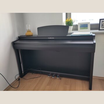Černý digitální piano s nápisem YAMAHA na přední straně. Nástroj má uzavřenou klaviaturu, tři pedály a na horní desce je umístěn notový stojan. Povrc…