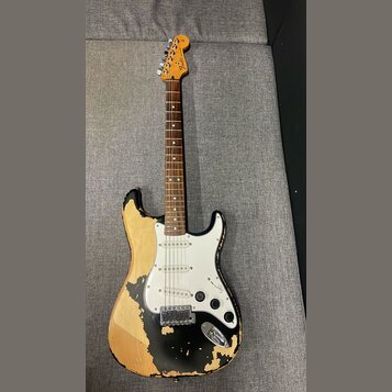 Elektrická kytara typu Stratocaster s černým lakem, který je výrazně odloupaný a odhaluje světlé dřevo korpusu. Na hlavě hmatníku je nápis Fender. Ky…