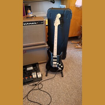Černá elektrická kytara typu Stratocaster s bílým hmatníkem a bílým pickguardem, opřená o černý tvrdý kufřík. Na hlavě kytary je nápis Fender STRATOC…