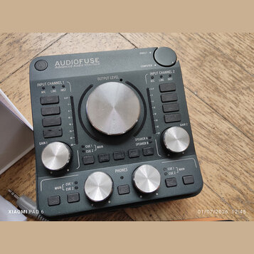 Tmavě šedý audio rozhraní s nápisem AUDIOFUSE Advanced Audio Interface. Zařízení obsahuje několik stříbrných otočných ovladačů s popisky GAIN 1, OUTP…