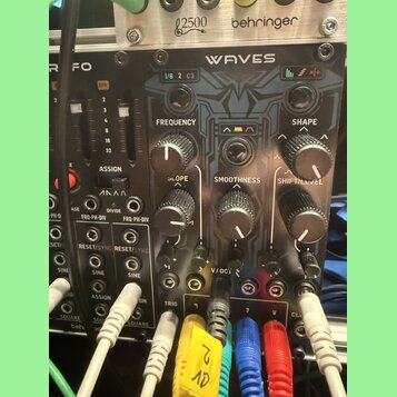 Černý modulární syntetizátor s nápisem WAVES. Na panelu jsou ovládací prvky s popisky FREQUENCY, SLOPE, SMOOTHNESS, SHAPE a SHIFT/LEVEL, dále značení…