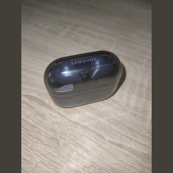 Černý nabíjecí box s průhledným víkem, uvnitř kterého jsou umístěna dvě bezdrátová sluchátka. Na horní části víka je bílý nápis SAMSUNG. Povrch průhl…