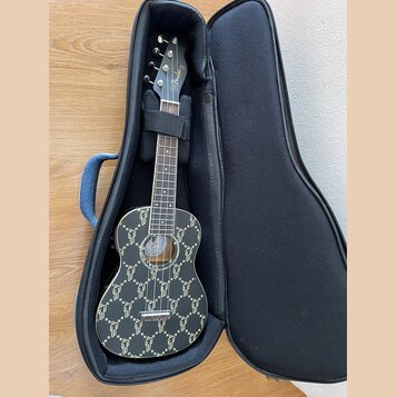 Černý ukulele v černém měkkém pouzdře. Tělo nástroje je pokryto celoplošným zlatým vzorem připomínajícím loga módních značek. Na hlavě je bílý nápis…