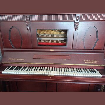 Hnědé dřevěné piano s klaviaturou a dekorativními výřezy na horním panelu. Na levé straně nad klávesami je nápis HUPFELD'S PHONOLISZT a na pravé stra…