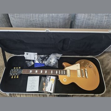 V otevřeném kufru leží elektrická kytara typu Les Paul v zlatavé barvě s krémovým pickguardem. Na hlavě kytary je nápis Gibson a modelové označení De…