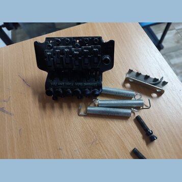 Černý kovový kobylník s nápisem Licensed under Floyd Rose Pats. Vedle něj leží tři kovové pružiny, jedna kovová opěrka a dva černé šrouby. Všechny dí…