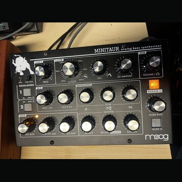 Černý analogový syntetizátor s nápisem MINITAUR analog bass synthesizer a logem Moog v levém dolním rohu. Zařízení má kovové tělo s mnoha stříbrnými…