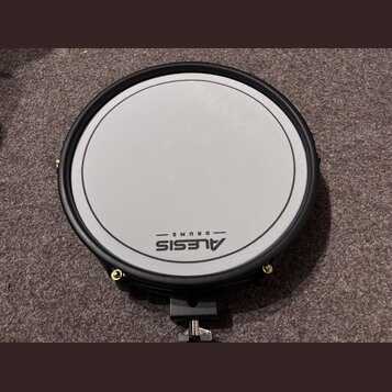 Kruhová elektronická bubenová pad v černé barvě s bílou úderovou plochou. Na spodní části plochy je černý nápis ALESIS DRUMS. Pad má černý obrubník a…