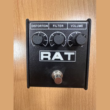 Černý kytarový efekt pedál s nápisem RAT v bílém rámečku. Horní část obsahuje tři černé otočné knoflíky s popisky DISTORTION, FILTER a VOLUME. V doln…