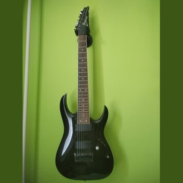 Černá elektrická kytara s tmavým hmatníkem, zavěšená na zelené stěně. Na hlavě nástroje je bílý nápis Ibanez. Kytara má dva humbuckery, jeden ovládac…