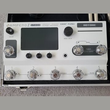 Bílý efektový procesor Hotone Ampero II Stage v hliníkovém kufru. Na horním panelu jsou nápisy AMPERO II STAGE, CDCM HD Gen 2 Tri-Core DSP Amp Modele…