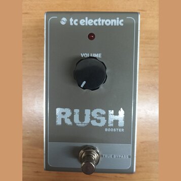 Stříbrný efekt pedál s nápisy RUSH a BOOSTER. Na pravé straně je logo tc electronic, vprostřed černý ovládací knoflík s popiskem VOLUME a červená kon…
