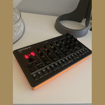 Černý sekvenční syntetizátor s oranžovými bočními panely. Na zařízení je bílý nápis Roland a model T-8, červený digitální displej zobrazující znaki „…