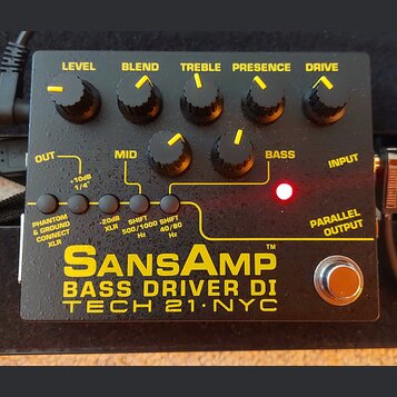 Černý basový předzesilovač s žlutými nápisy a ovládacími knoflíky. Na zařízení jsou uvedeny názvy SansAmp, BASS DRIVER DI a TECH 21 NYC. Horní řada k…