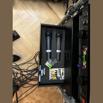 Otevřený černý kufřík s pěnovou vložkou, ve kterém jsou umístěny dva černé bezdrátové mikrofony. Na jednom z nich je vidět bílý nápis „SENNHEISER“ a…