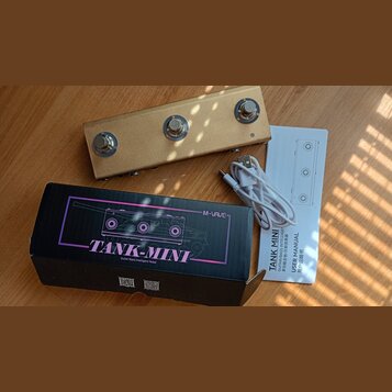 Fotografie zobrazuje zlatý efekt pedál s třemi stoupacími spínači, černou krabici a bílý uživatelský manuál s přiloženým USB kabelem. Na krabici je f…