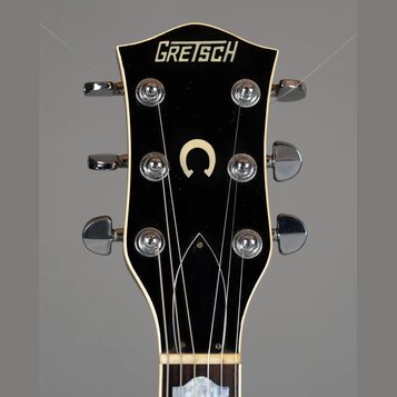 Detail hlavy elektrické kytary značky GRETSCH v černé barvě s bílým lemováním. Uprostřed hlavy je umístěno bílé písmeno C. Jsou vidět šest chromovaný…
